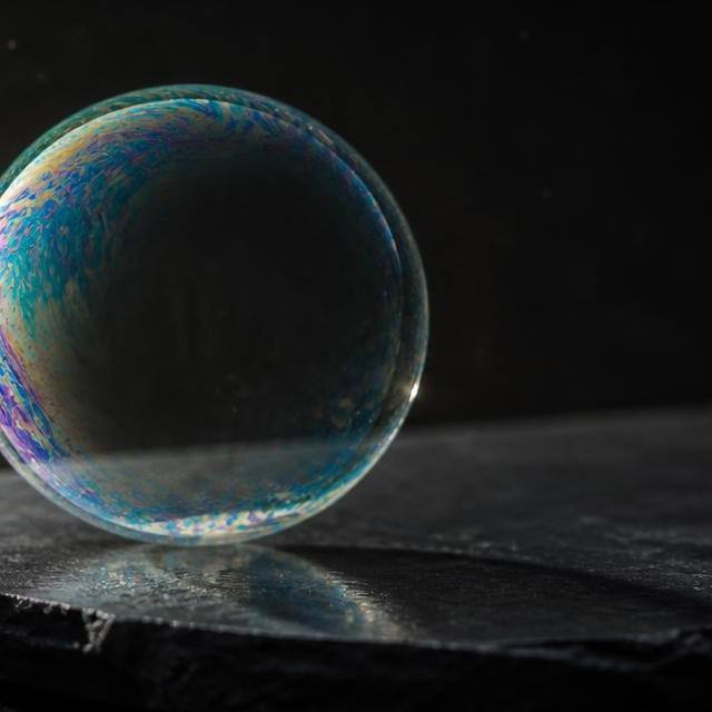 A single iridescent soap bubble resting intact on dark slate under a hard raking light