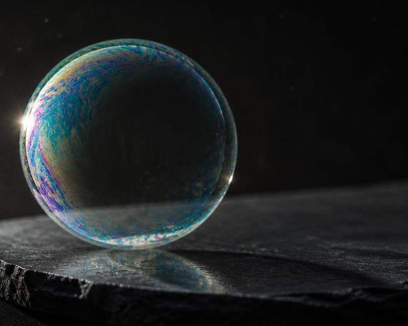 A single iridescent soap bubble resting intact on dark slate under a hard raking light